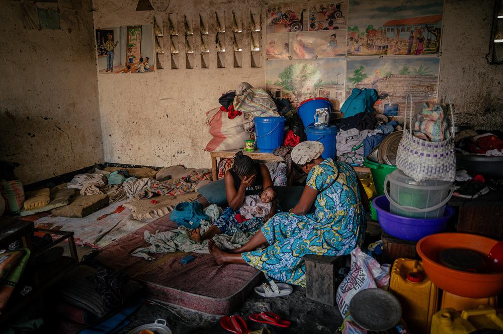 Deux femmes et leurs enfants, sont assis à l’extérieur d’une école avec leurs matelas, vêtements et autres effets personnels. Elles se sont réfugiées dans l’établissement scolaire après avoir fui les violences, et doivent sortir leurs affaires chaque matin pour laisser la place aux élèves pendant les cours. Leurs maris, militaires, ont disparu lors de l’offensive des M23 sur la ville de Goma en janvier et février 2025. 