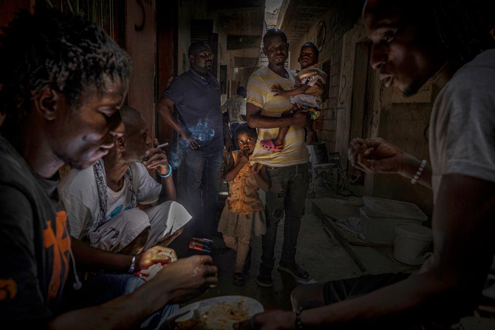 A group of migrants and refugees eat and smoke while a father and his two children watch them. Libya, August 2021.  