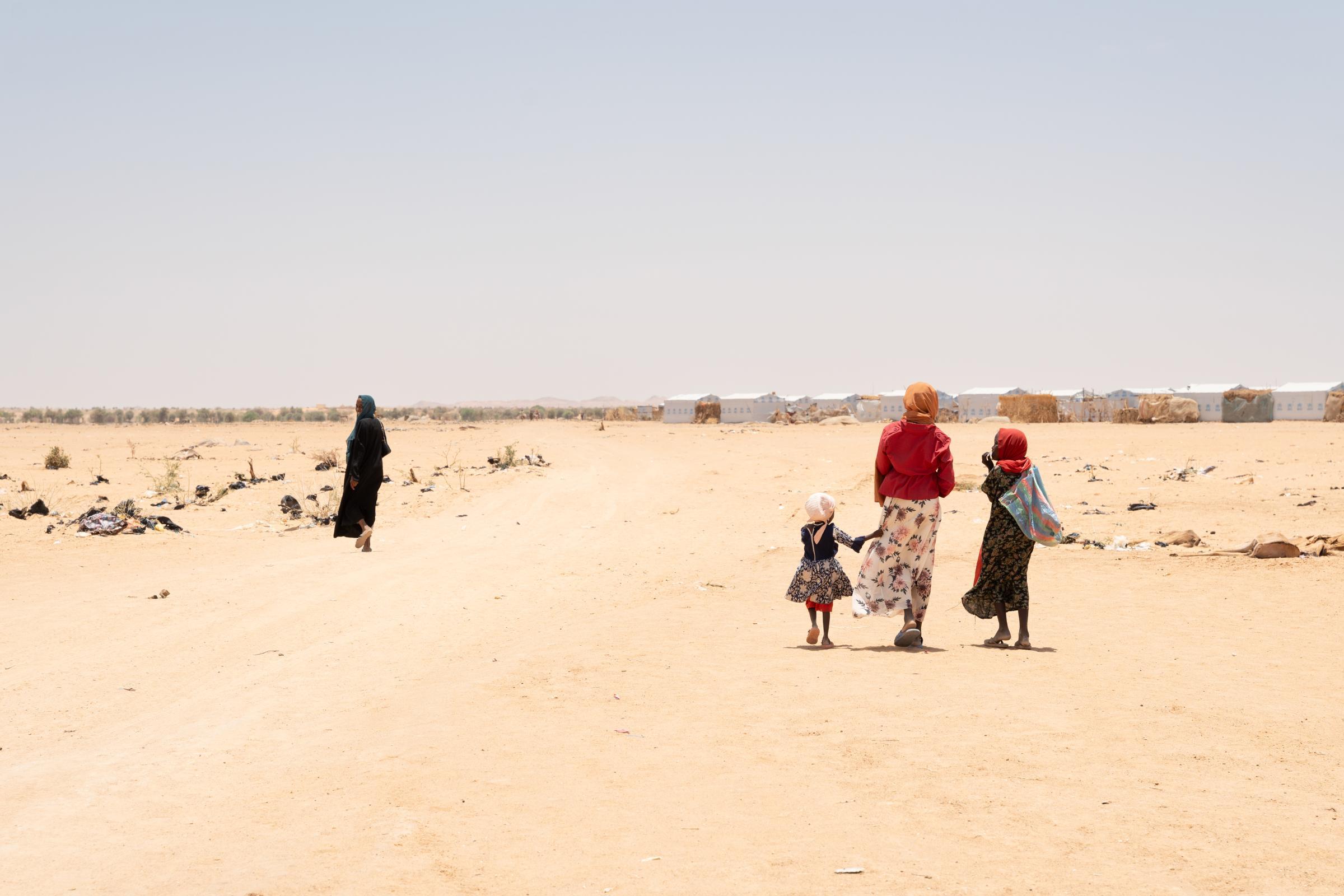 Des femmes et des enfants marchent sur un chemin de sable en direction des abris de la zone E du camp d'Ouré-Cassoni, Tchad. Le camp est situé dans une zone plate et aride du Sahel, où l’ombre est quasi inexistante et les températures élevées.