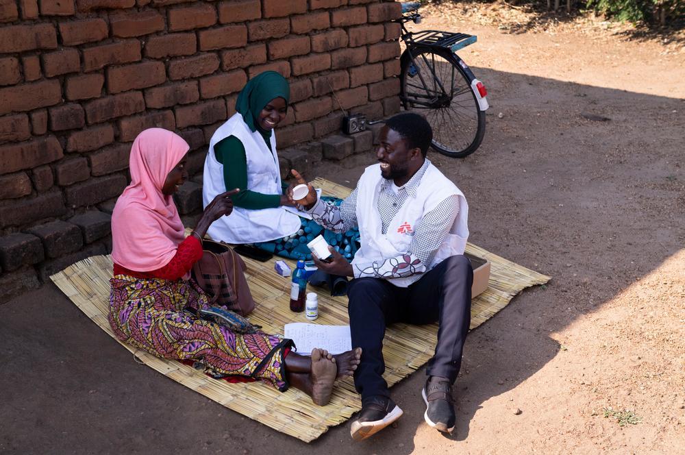 Deux membres de l'équipe de soins palliatifs de MSF, Wilfred Nkhambule et Vitu Mwanza, infirmiers en soins palliatifs, passent en revue les médicaments de Jean Gama lors d'une visite à domicile à Nkando, dans le district de Mulanje, au Malawi. L'équipe lui a appris à s'administrer elle-même ses traitements et lui retire ceux qu'elle tolère mal. Juillet 2026 © Jérémy Lempin